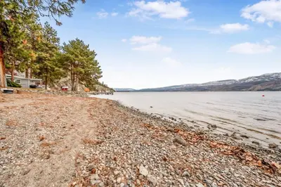 a rocky shore of a lake with a house and trees at Secluded Cabin On The Lake #7 in West Kelowna