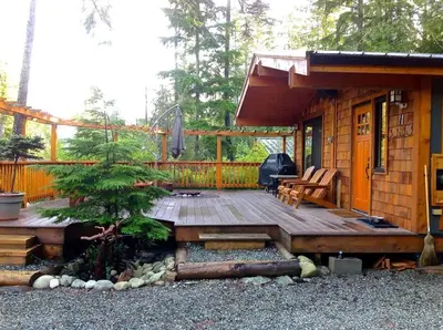 a small cabin with a wooden deck and a tree at French Girl's Lair - Pet Friendly - Garden View in Port Renfrew