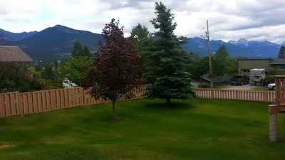two trees in a yard with a fence and mountains at Invermere 4 Br Lakeside, Mountain View Home! Walk to Sandy Beach for Swimming, Boating, Tennis and Beach Volleyball Local Town Shopping, Eateries, Bakery and Pubs! Welcome Family's, Healthcare Workers and Trades! Monthly Rentals of 28 Nights or More! in Invermere