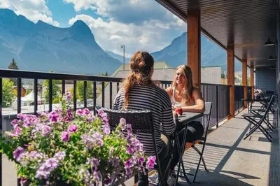 two women sitting on a balcony with mountains in the background at Lamphouse By Basecamp in Canmore