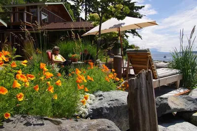 a woman sitting in a garden with an umbrella and flowers at Blitz Beach House Oceanside Suite in Powell River