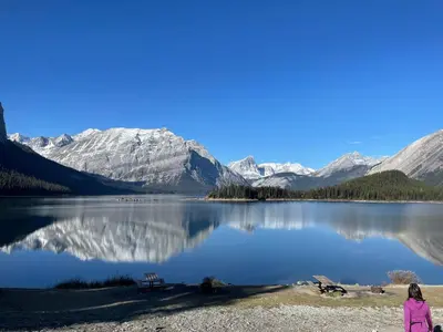 a woman sitting on the shore of a lake with snow covered mountains at Rundle Rise - Spacious Condo Steps from Main Street in Canmore
