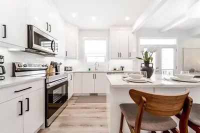 a white kitchen with white cabinets and a wooden chair at Modern Nordic Escape, 3BR Near WEM & DT, AirCon & Fireplace, Wifi, Sleeps 6! in Edmonton